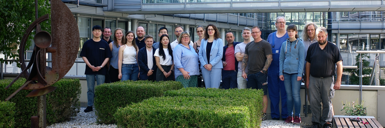 Mitglieder der Arbeitsgruppe Alloy Design and Processing vor einem modernen Glasgebäude stehend, einige mit lockerer, sommerlicher Kleidung, andere mit Hemden und Blazern.