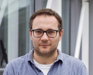 Anatol Prudnikau with short brown hair wearing a gray shirt over  a white t-shirt standing in front of a blurred background with glass  and metal structures.