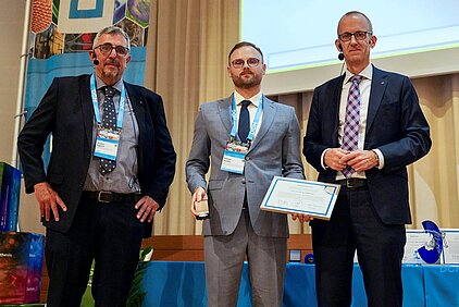 Three men in suits stand side by side, the man in the middle is Dr. Konrad Kosiba holding a certificate and an award, in a conference room with a projection screen in the background.