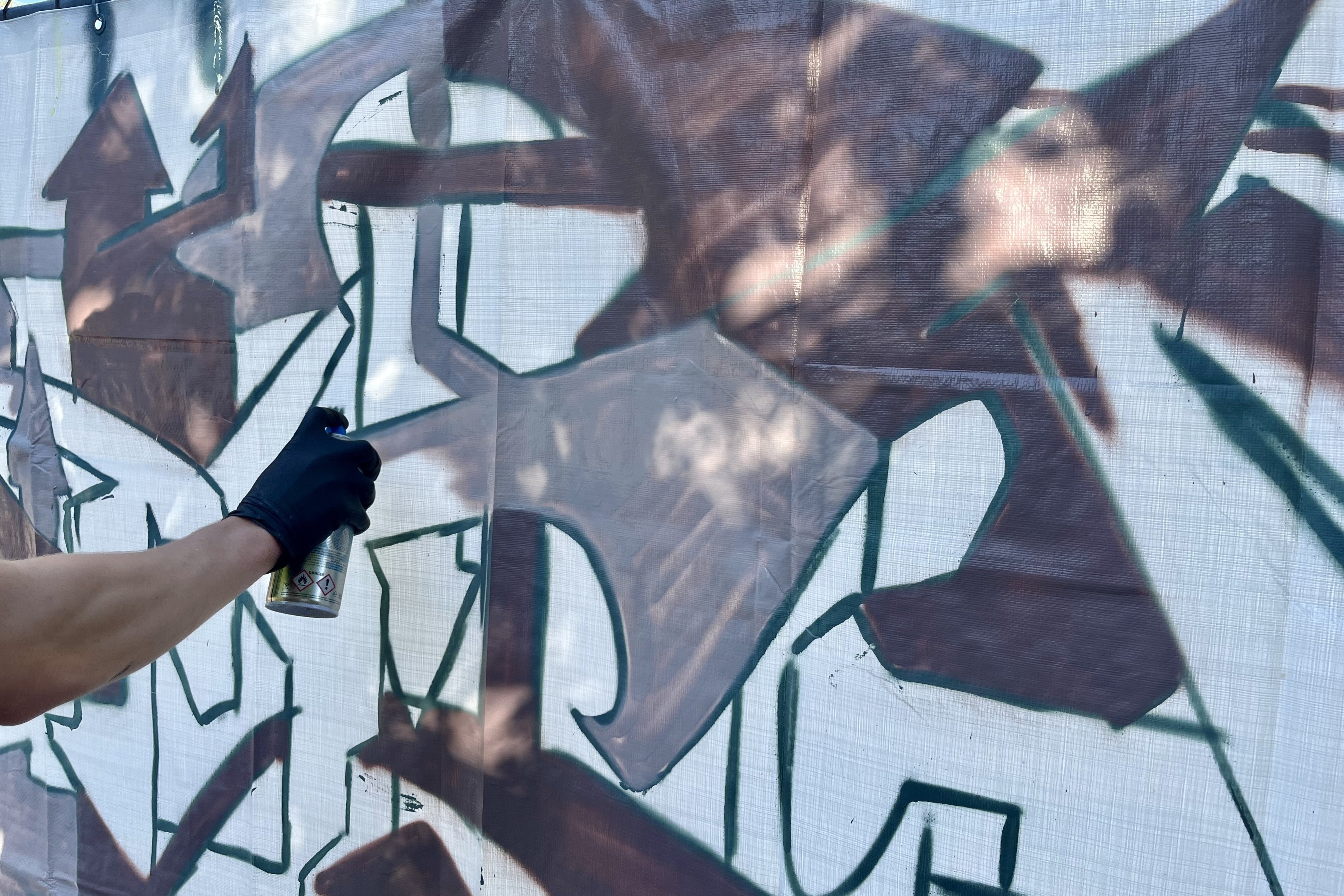 Eine Hand mit schwarzem Gummihandschuh und Sprayflasche. Es entsteht ein Graffiti in gäulichen Farben auf der Bespannung eines Bauzaunes.. 