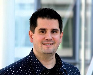 Dr. Fabian Paulus with brown hair wearing a plaid shirt over a white  T-shirt against a plain background. 