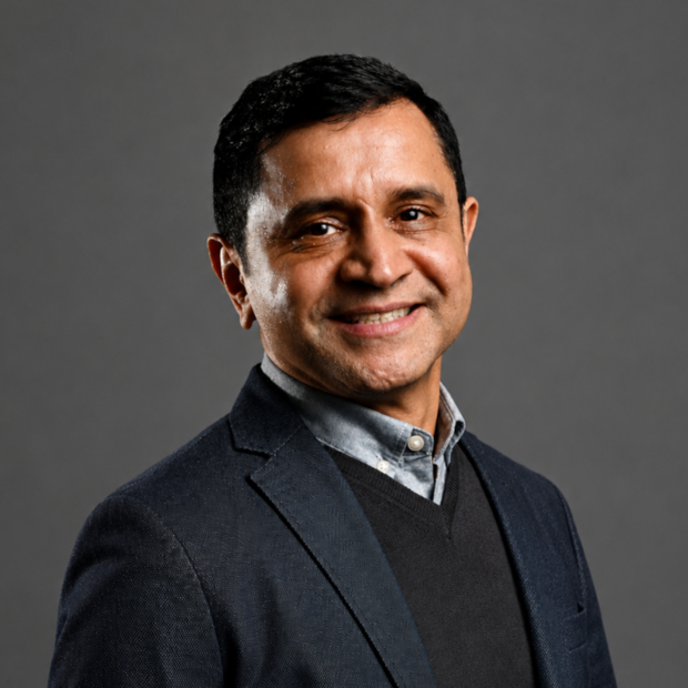 Dr. Harish Parala with dark hair wearing a gray blazer, light blue shirt, and dark sweater against a gray background, face blurred.