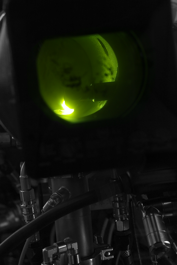 View through a round observation window into a green-lit chamber with metallic components and connected hoses and valves.
