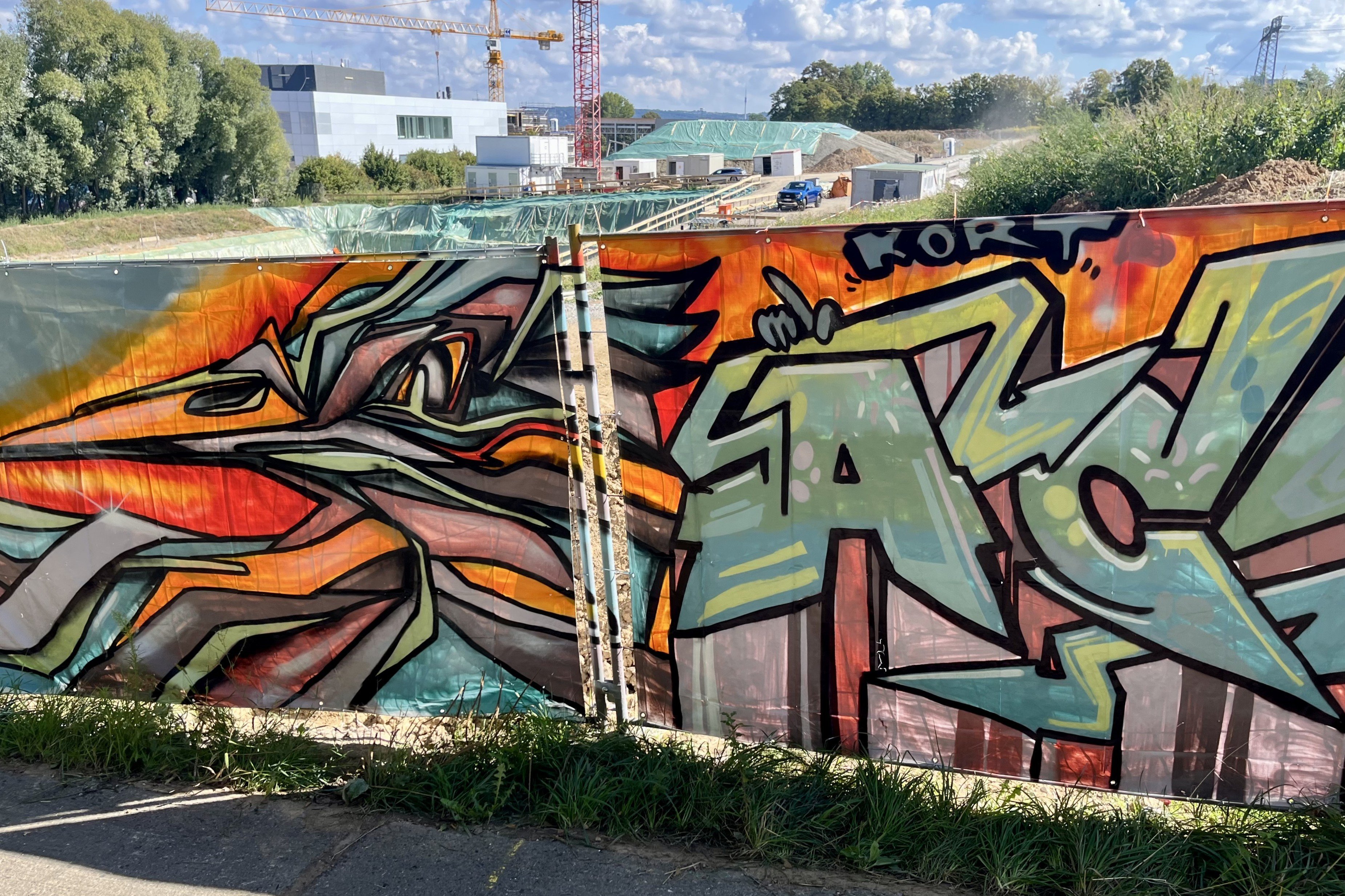 Graffitikunst am Bauzaun in den Farben orange, rot, türkis. Ein bunter Schriftzug. Im Hintergrund ist die Baugrube des künftigen Forschungsgebäudes zu sehen.