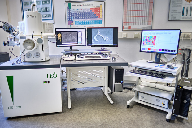 Laboratory setup with an electron microscope, two monitors on a desk, and another monitor on a rolling cart in a room with scientific posters on the wall.