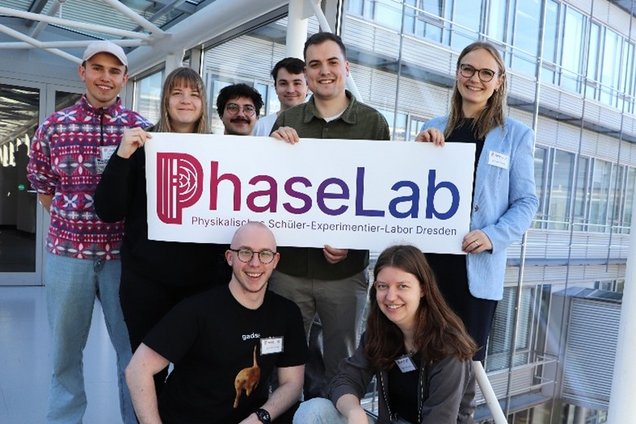 Group of eight people holding a sign reading 'PhaseLab Physikalisches Schüler-Experimentier-Labor Dresden' in front of a modern glass building.