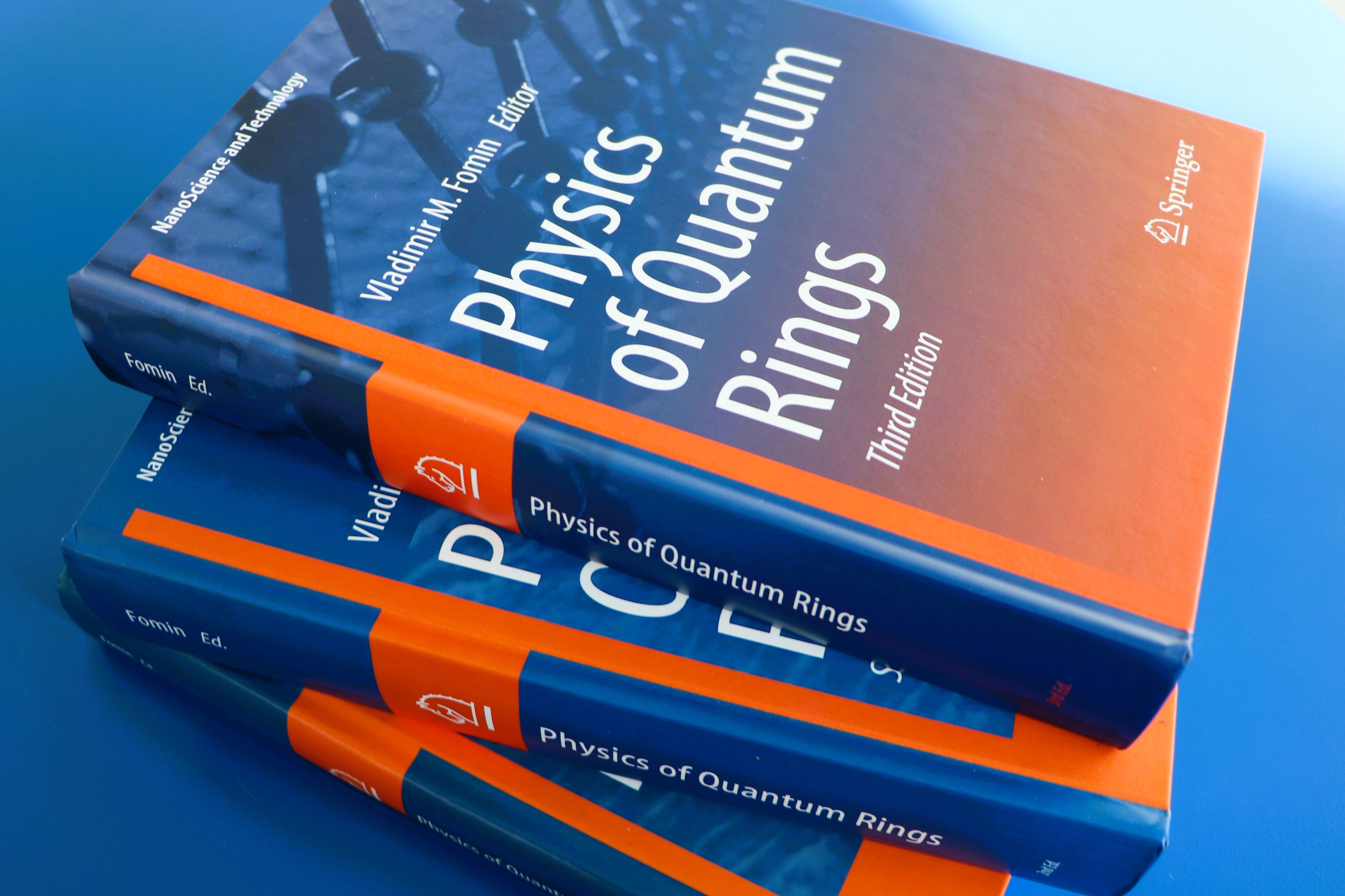 Drei Bücher liegen auf einem Stapel. Das oberste Buch ist die 3.Auflage mit dem Titel "Physics of Quantum Rings". Das Buch ist orange-blau gestaltet.