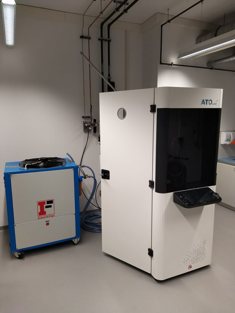 Metal powder atomization system ATO Lab Plus consisting of a white rectangular main unit with a black front door and a blue cubic auxiliary unit with cables in a laboratory room.