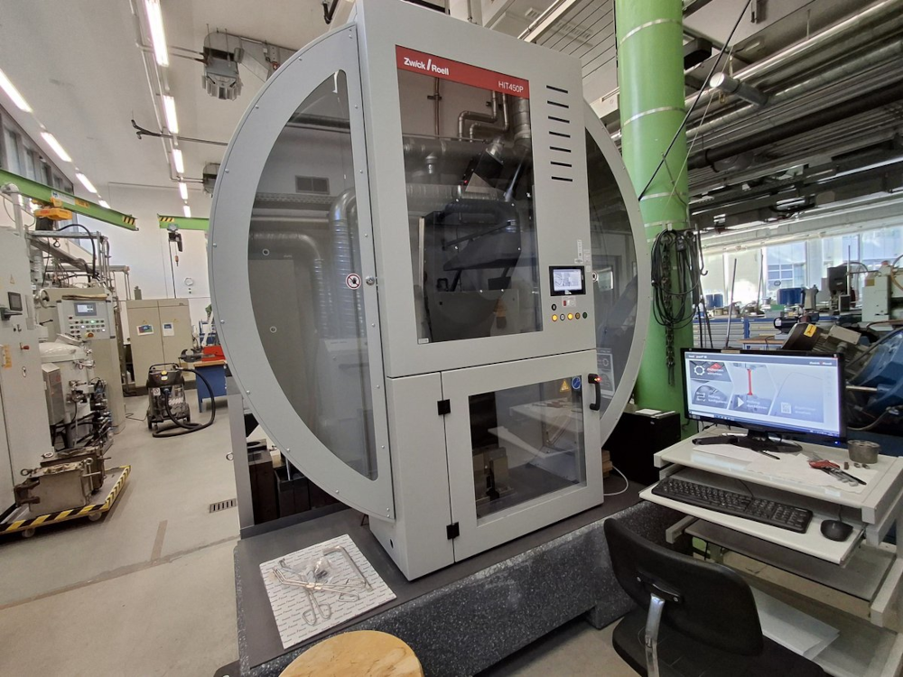 A Zwick HIT 450P impact bending tester with a round front window in a laboratory, with a computer workstation next to it.