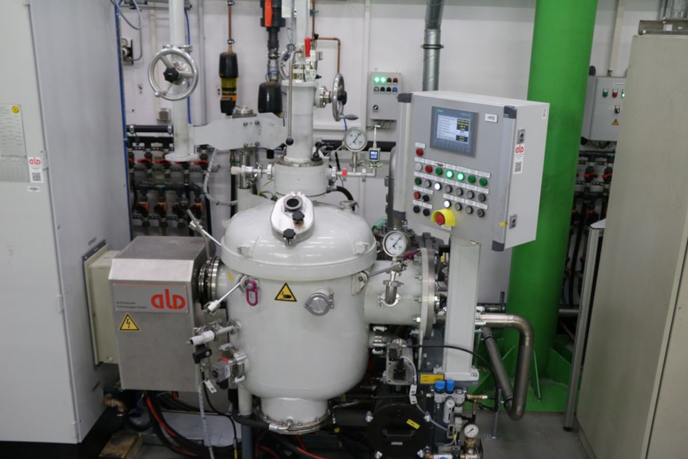 Induction melting system with a round vessel, control panel featuring a display and multiple buttons, and various pipes and gauges in a technical room.