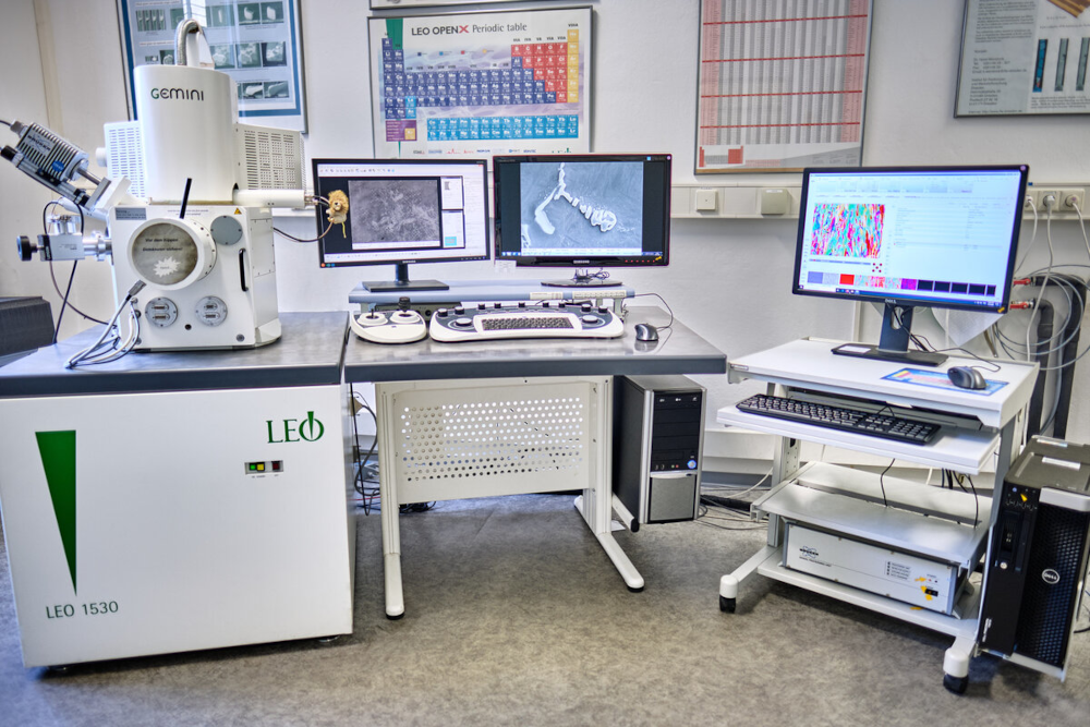 Laboratory setup with an electron microscope, two monitors on a desk, and another monitor on a rolling cart in a room with scientific posters on the wall.