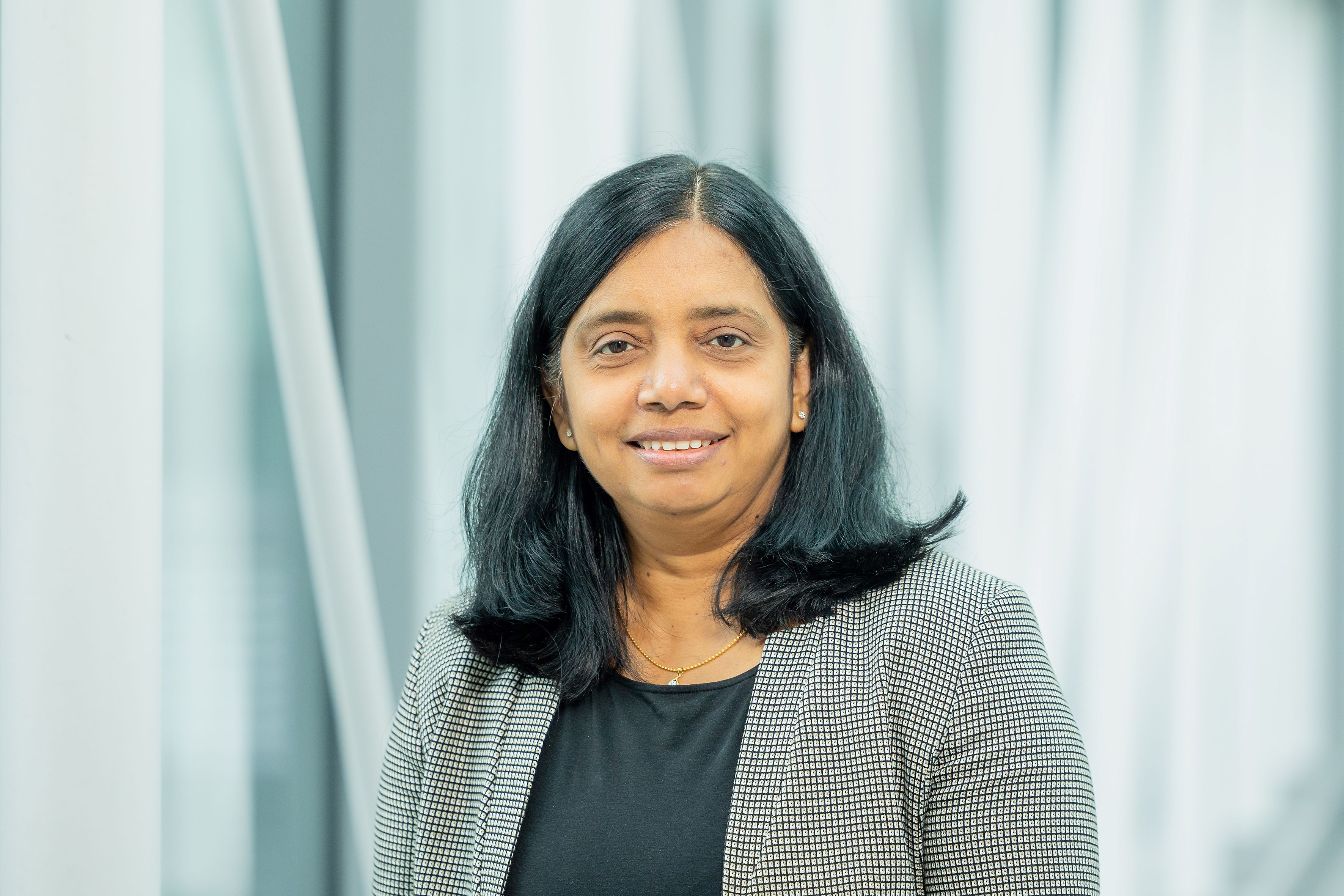 Prof. Dr. Anjani Devi with shoulder-length straight hair wearing a checkered blazer over a black top standing in front of a modern light background with glass windows at IFW Dresden.
