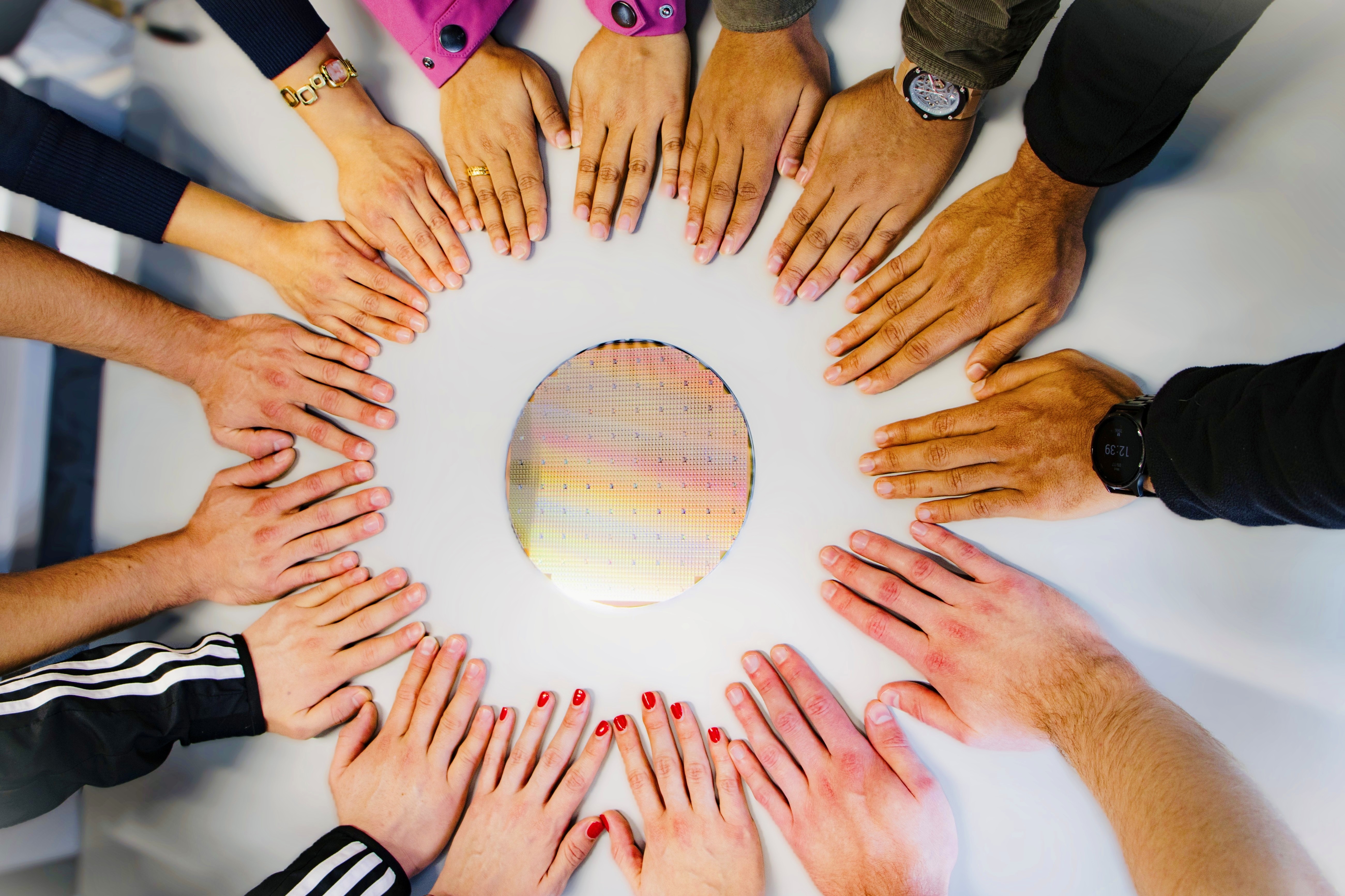 Many hands of different skin colors form a circle around a round, shiny wafer.