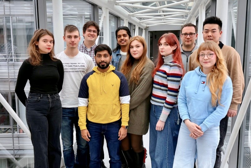 Group of nine scientist of the Team “Sustainable Nanomaterials” standing in a bright room with glass windows and visible steel beams in the background.