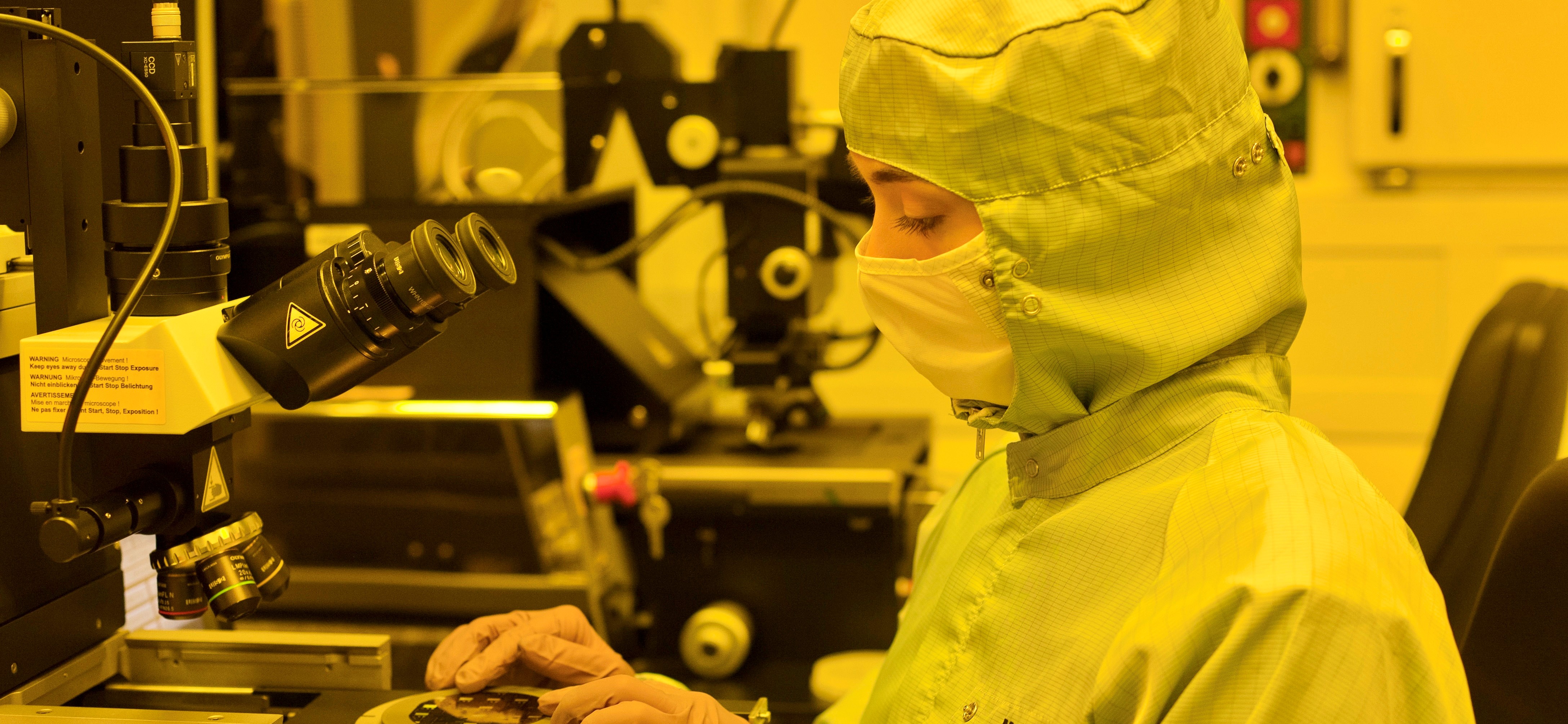 Person in protective clothing and face mask working at a microscope in a laboratory with yellow lighting.